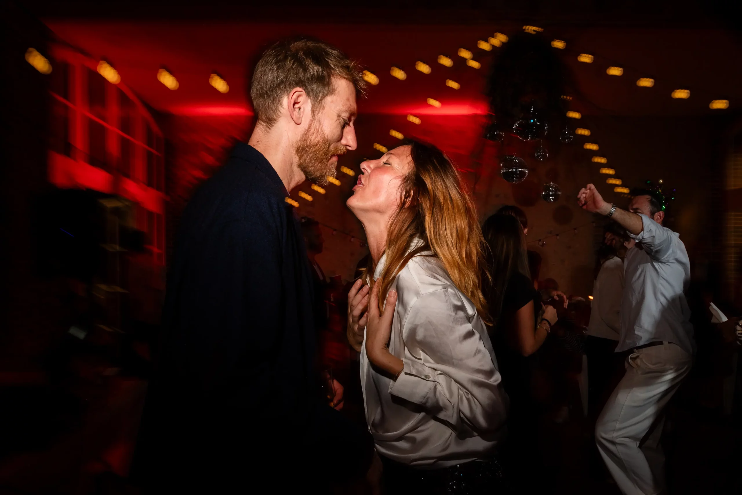 mariage-angers-bal reportage documentaire de mariage, photo prise pendant une soirée, en premier plan un couple se fait face. la femme chante et l'homme la regarde tendrement. en arrière plan on voit le jolie dispositif lumineux et la lumière du flash rouge qui permet de donner une ambiance festive