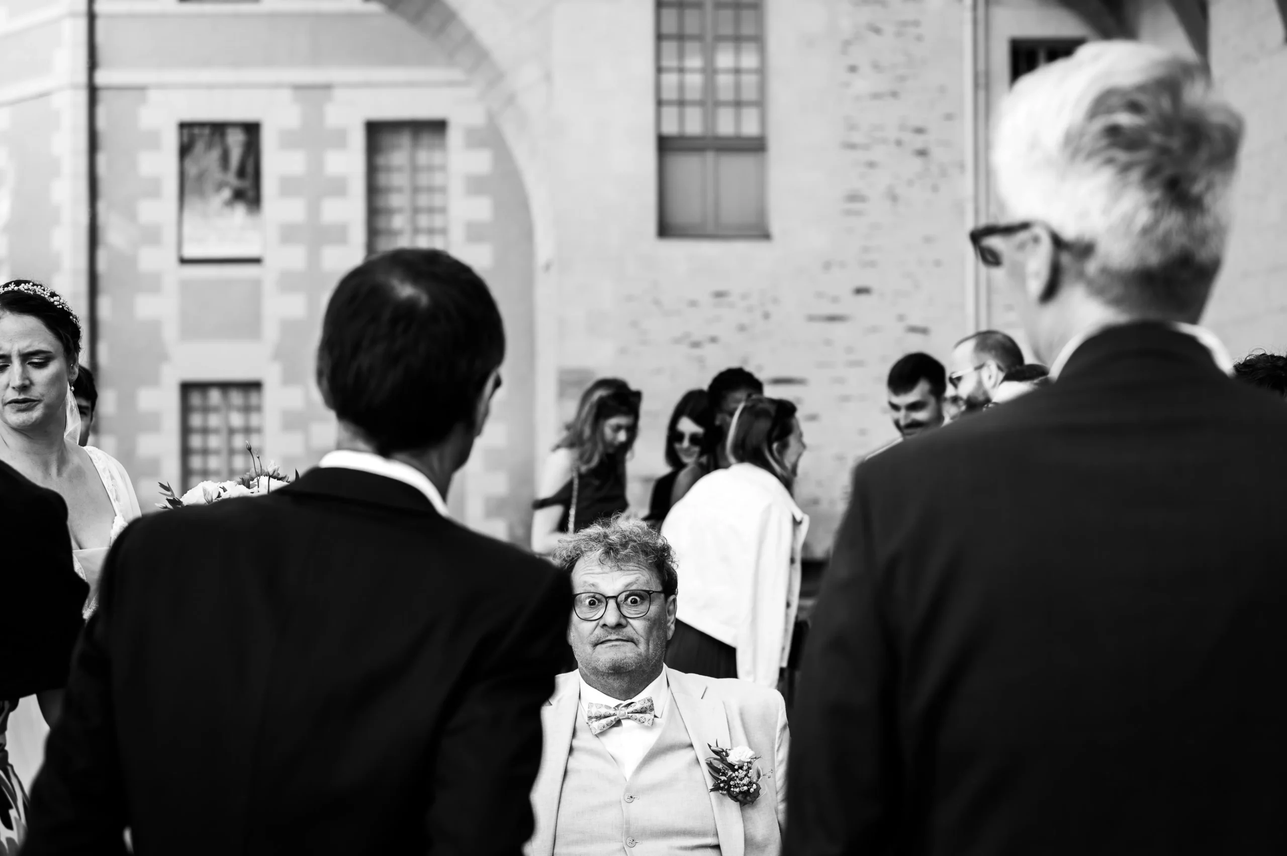 photographe-mariage-mairie-nantes photographie de mariage à la sortie de la mairie de Nantes. Photographie en noir et blanc, avec en premier plan le papa de la mariée assis sur une chaise, qui me regard avec de gros yeux. photographie drôle